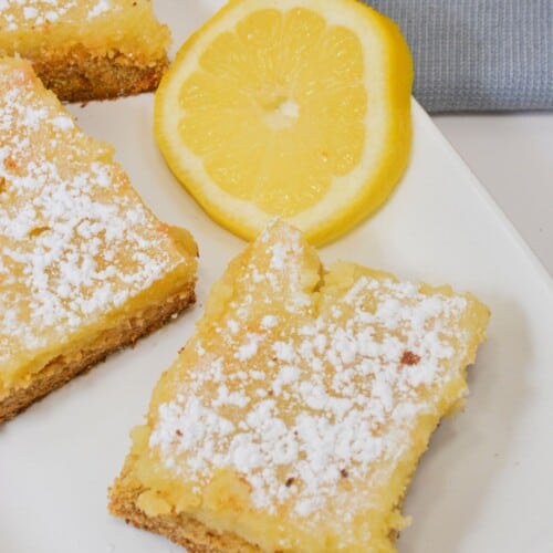 Two lemon bars on a plate with a slice of lemon on the side.